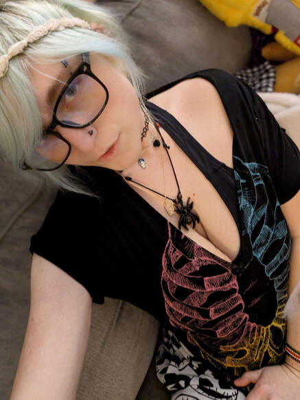 Picture of Myrtle with lightly tinted blue-white hair wearing black square glasses and a white flower crown. Their shirt is black with a rainbow ribcage. Their necklace is of a large brown spider, and are wearing a basic black tattoo stretch choker.